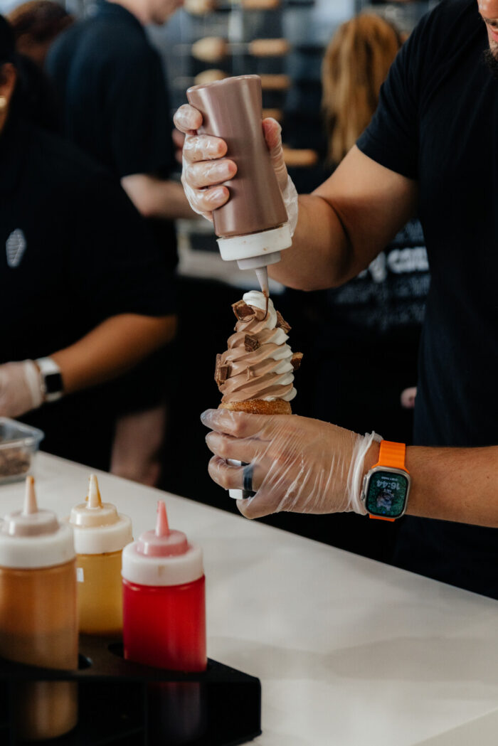 A person wearing gloves and an orange smartwatch squeezes chocolate syrup onto a swirled ice cream cone. Various sauce bottles are on the counter. People work in the background.