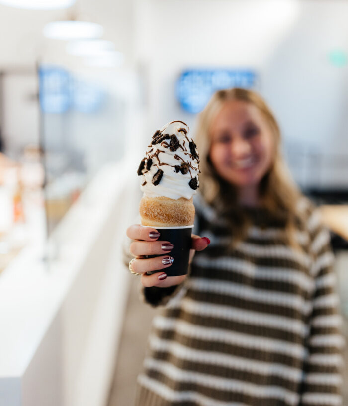 A woman holding a large soft serve ice cream cone with chocolate drizzle and crushed toppings, smiling in a brightly lit indoor setting. The ice cream is in focus, while the woman is blurred in the background.