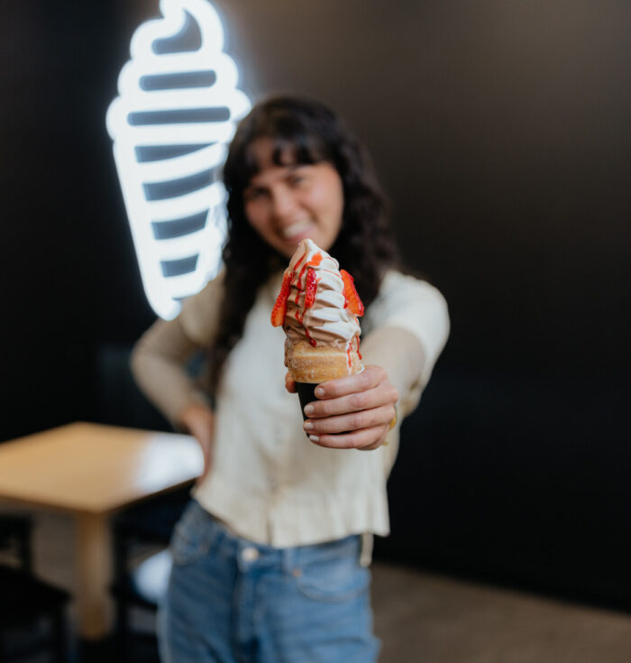 A woman holds a soft-serve ice cream cone with strawberry topping toward the camera, smiling, with a neon ice cream sign in the blurred background.