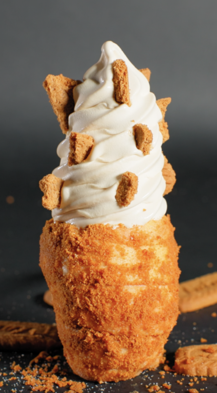 A tall swirl of vanilla soft serve ice cream in a crispy, crumb-coated cone, topped with pieces of cookie, against a dark background. Cookie crumbs and pieces are scattered around the base.