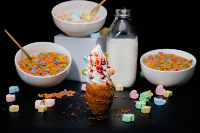 Soft serve ice cream topped with red syrup and colorful cereal pieces in a cone, surrounded by bowls of cereal, a bottle of milk, and scattered marshmallows on a black background.