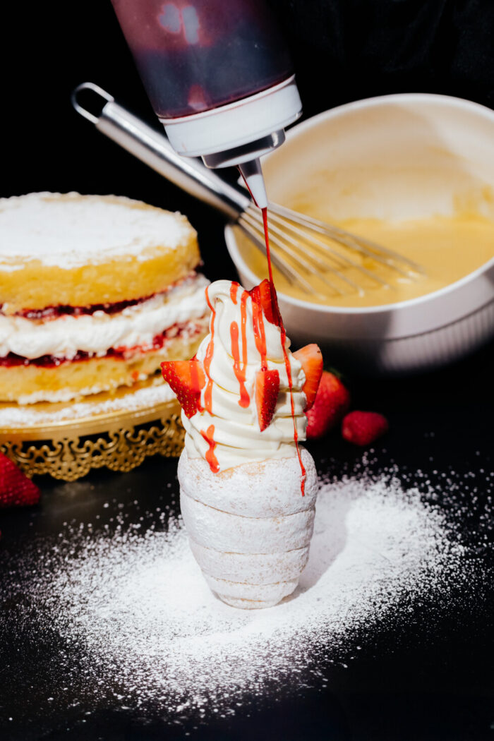 A hand pours red syrup over soft serve ice cream topped with sliced strawberries, served in a pastry cone. In the background, there’s a layered sponge cake with cream and a mixing bowl with a whisk, all dusted with powdered sugar.