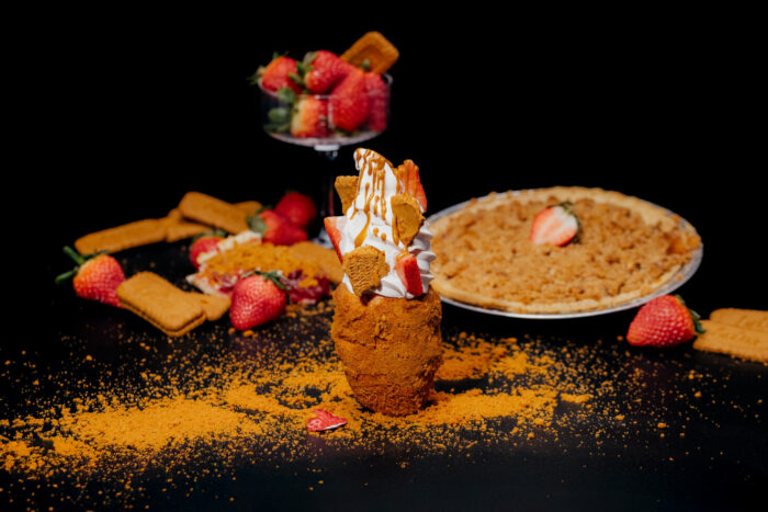 A dessert in a crumb-coated glass is topped with whipped cream, strawberries, caramel drizzle, and biscuit pieces. Surrounding it are strawberries, biscuits, a bowl of berries, and a crumb pie, all on a black background.