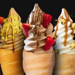 Five soft serve ice creams in pastry cones, topped with various sauces, strawberries, cookie pieces, and nuts, displayed side by side against a black background.