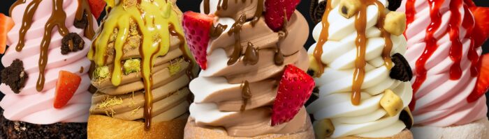 Five soft serve ice creams in pastry cones, topped with various sauces, strawberries, cookie pieces, and nuts, displayed side by side against a black background.