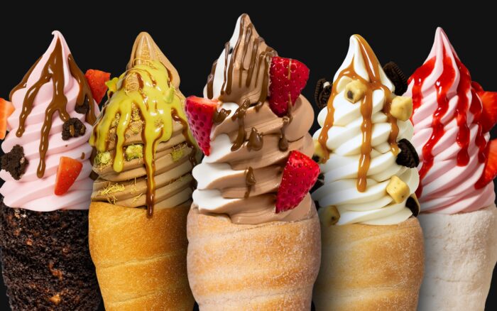 Five soft serve ice creams in pastry cones, topped with various sauces, strawberries, cookie pieces, and nuts, displayed side by side against a black background.