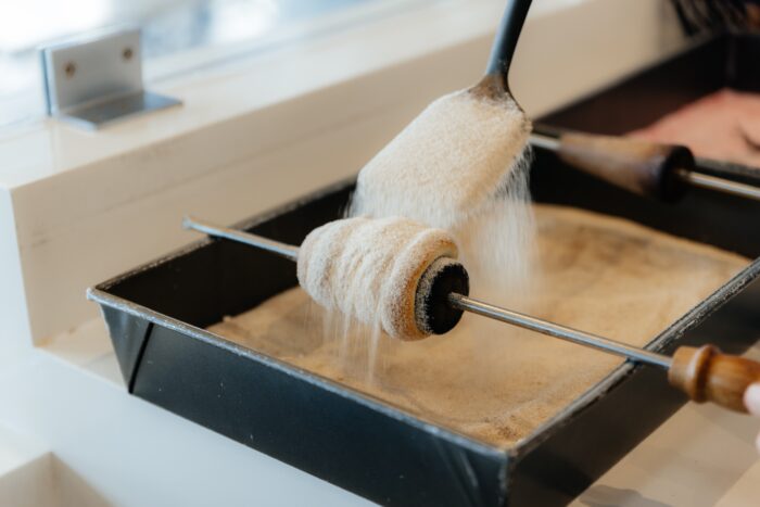A hand sprinkles sugar over a pastry wrapped around a metal spit, with the pastry resting above a tray filled with sugar. The scene takes place indoors, likely in a bakery or kitchen.