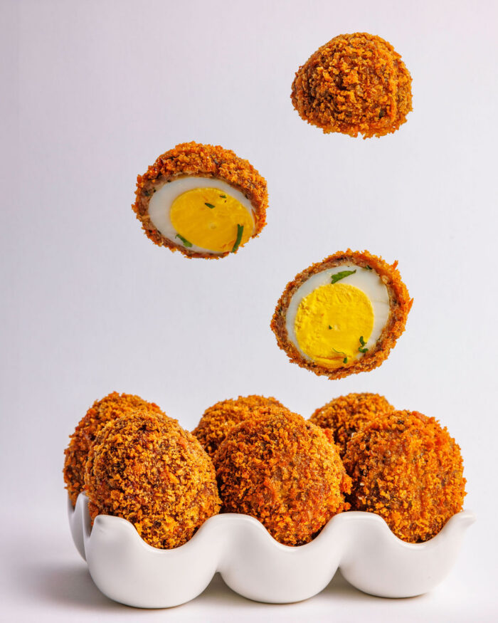 A white egg tray holds several crispy, breaded Scotch eggs crafted by Pastry Queens. Two Scotch eggs, one cut in half to reveal the yolk and herbs inside, appear to float above the tray against a plain background.