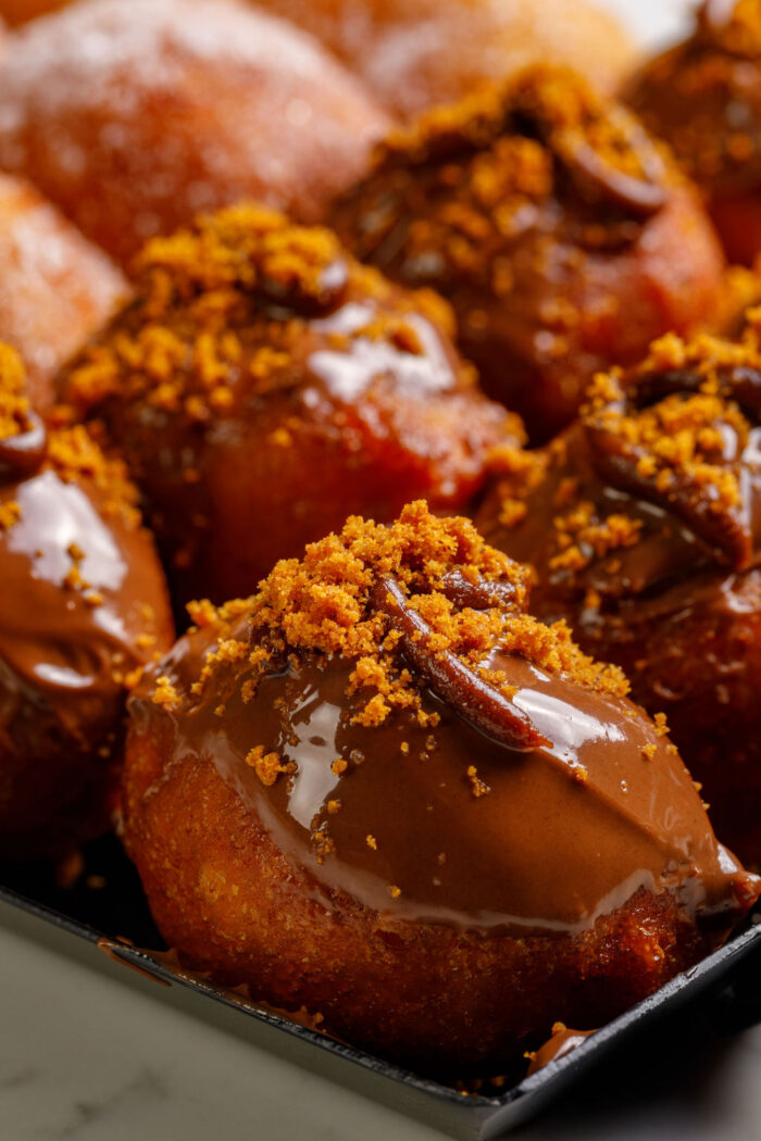 Close-up of Pastry Queens donuts topped with glossy chocolate glaze and sprinkled with crunchy brown crumbs, arranged closely together on a tray.