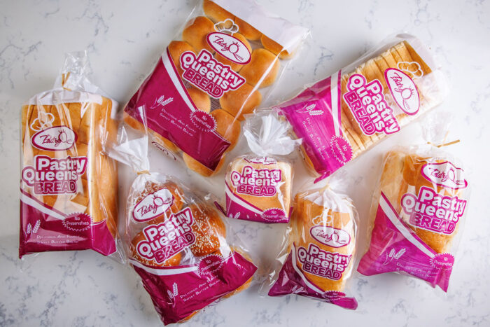 Seven loaves of Pastry Queens Bread in various packaging are arranged on a white marble surface. The bags—white, pink, and purple—each feature the Pastry Queens logo and product name for a colorful, inviting display.