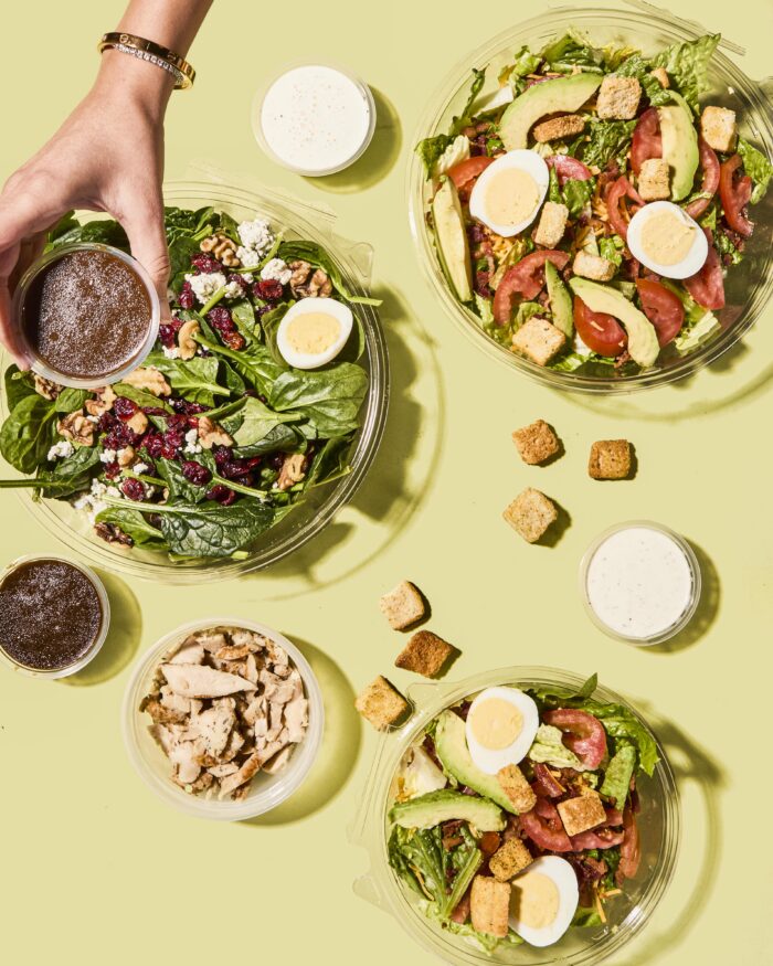 Three fresh salads with greens, eggs, avocado, walnuts, cranberries, tomatoes, and croutons are arranged on a yellow surface, along with dressings and a bowl of grilled chicken. A hand holds a cup of dressing over one salad.
