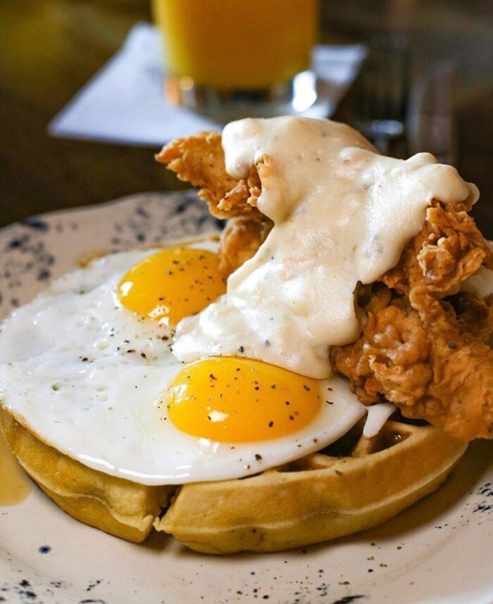 Haywire Brunch which is a plate with a waffle topped with two sunny-side-up eggs, crispy fried chicken, and creamy white gravy—perfect for breakfast lovers searching for tasty spots in places near DFW. A glass of orange juice is blurred in the background.
