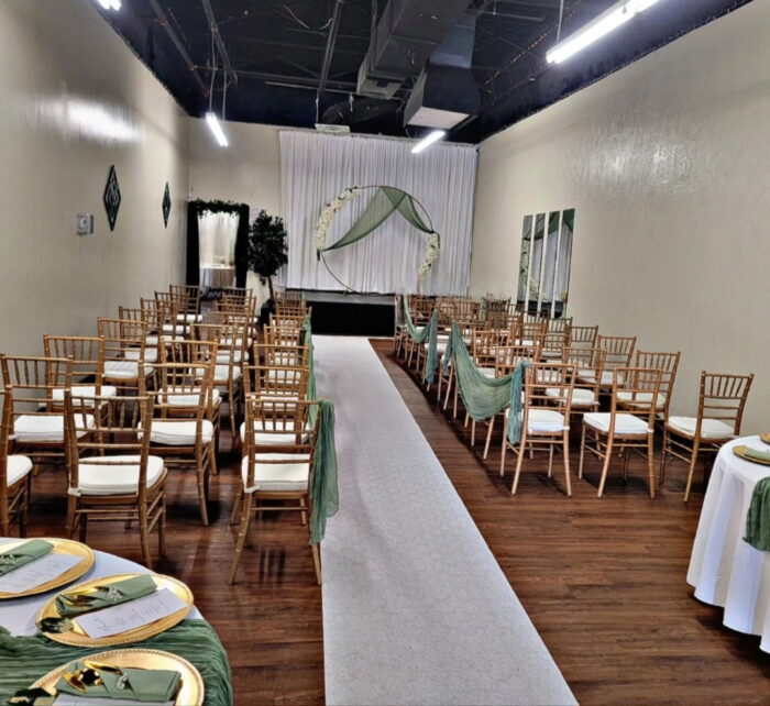 A wedding ceremony setup with rows of wooden chairs draped with green fabric, a white aisle runner, and a simple arch decorated with greenery at the front of the room. Tables with gold plates and green napkins are visible at venueiztic.
