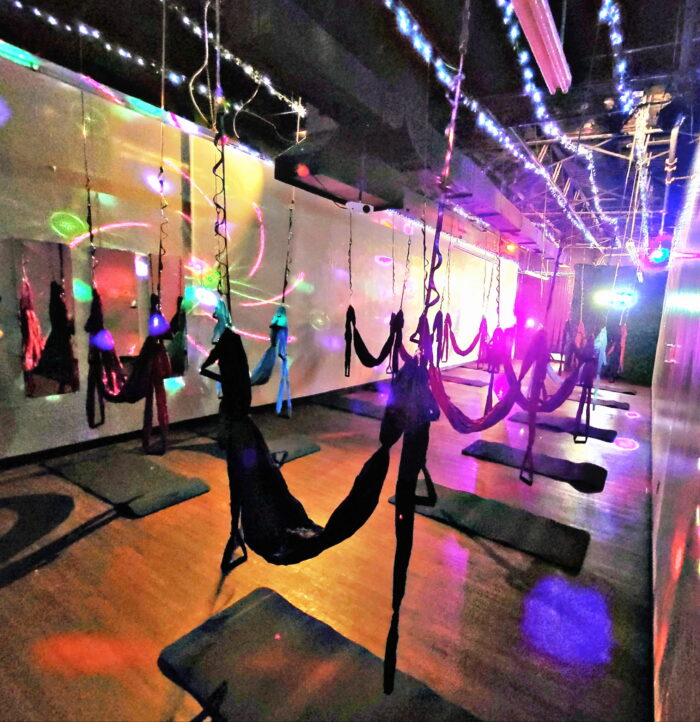 A dimly lit room with aerial yoga hammocks hanging from the ceiling, colorful lights and reflections on the walls and floor, with yoga mats placed beneath each hammock. The atmosphere is energetic and vibrant at venueiztic.