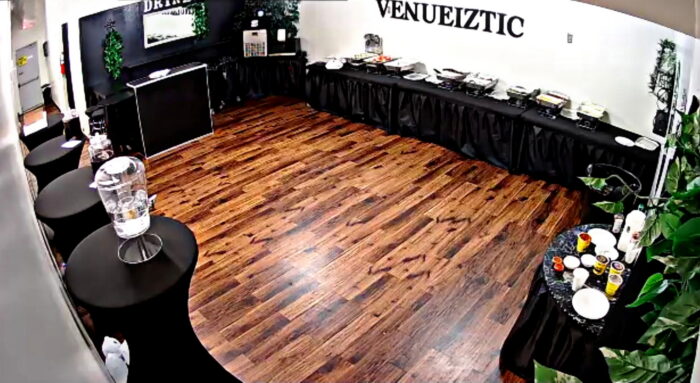 A banquet room with wooden floors, black-draped tables set with food trays and drinks, a black counter, and VENUEIZTIC written on the white wall. The space appears ready for an event at venueiztic.