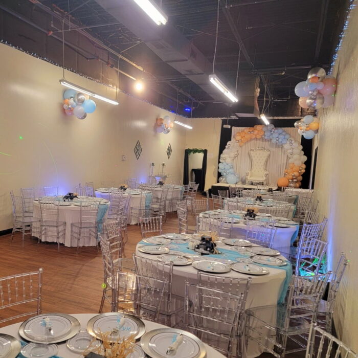 A decorated event hall with round tables covered in white tablecloths and blue accents, clear chairs, balloon arrangements, and a balloon arch framing a white backdrop at the end of the room at venueiztic.