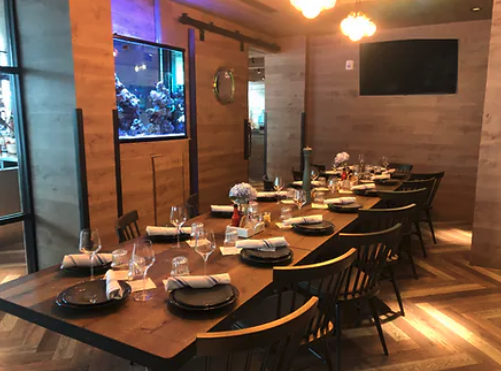 A modern restaurant dining area with a long wooden table set for a meal, black chairs, wine glasses, and neatly folded napkins at Urban Seafood Co.. Soft lighting and a large fish tank create a cozy atmosphere.