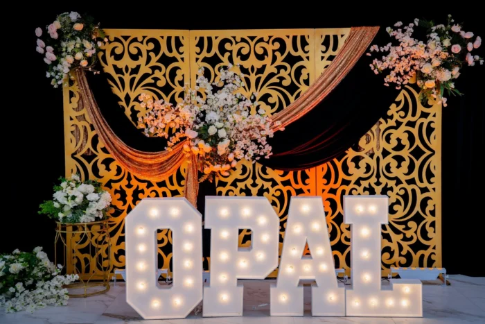 Large illuminated letters spelling OPAL are displayed in front of a decorative gold screen with floral arrangements and black and gold drapes, creating an elegant backdrop.