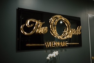A black sign with shiny gold cursive text reads The Opal VENUE mounted on a dark wall, with a small white flower arrangement visible at the bottom.