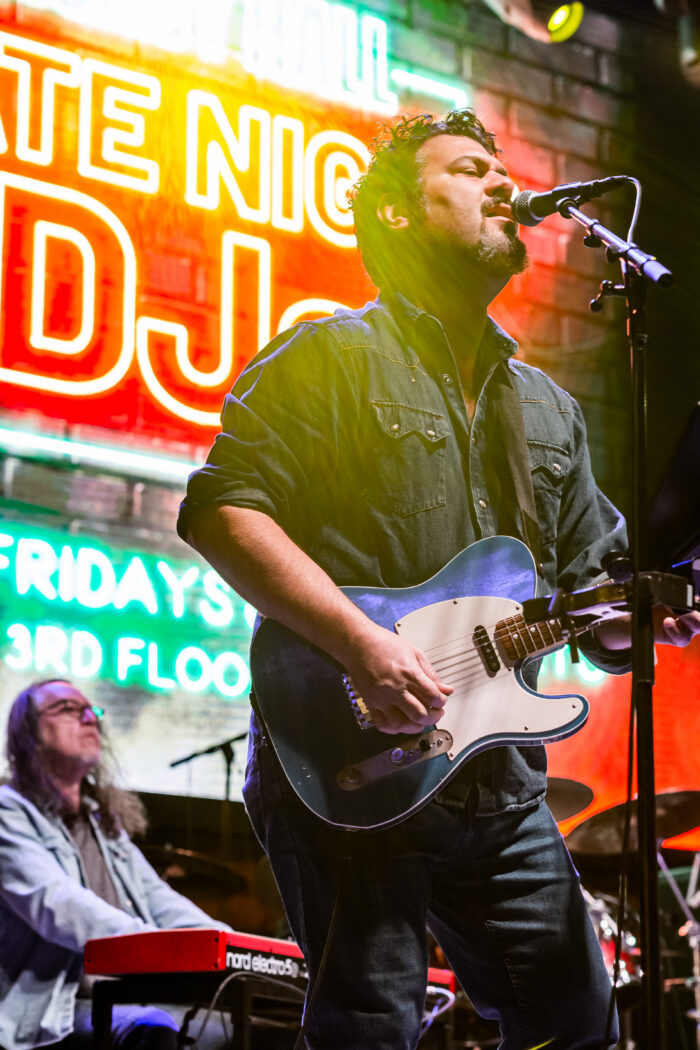 A musician plays an electric guitar and sings into a microphone on stage while another person plays a keyboard in the background, creating a lively atmosphere filled with Fun Things To Do. Bright neon signs with bold text are visible behind them.