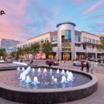 A modern outdoor plaza at sunset features illuminated water fountains, people walking, trees, and surrounding shops. The sky is pink and blue. The mindtrip. logo appears in the upper left corner.