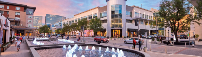 A modern outdoor plaza at sunset features illuminated water fountains, people walking, trees, and surrounding shops. The sky is pink and blue. The mindtrip. logo appears in the upper left corner.