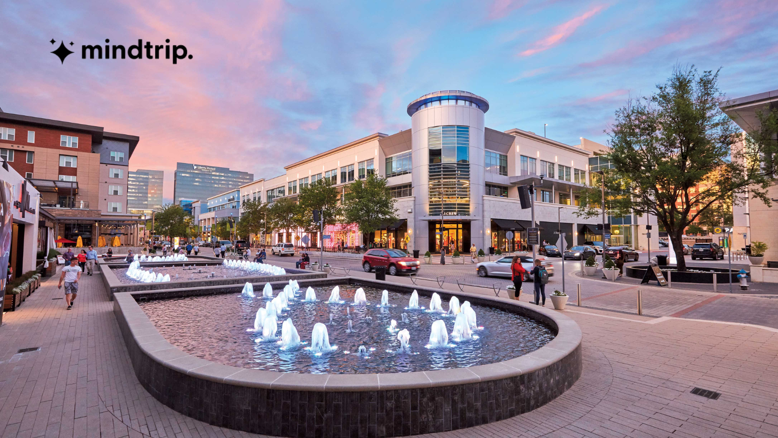 A modern outdoor plaza at sunset features illuminated water fountains, people walking, trees, and surrounding shops. The sky is pink and blue. The mindtrip. logo appears in the upper left corner.