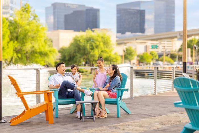 A smiling family of four relaxes on colorful Adirondack chairs on a wooden deck by the water—one of the best family vacation spots—surrounded by tall city buildings and green trees on a sunny day.