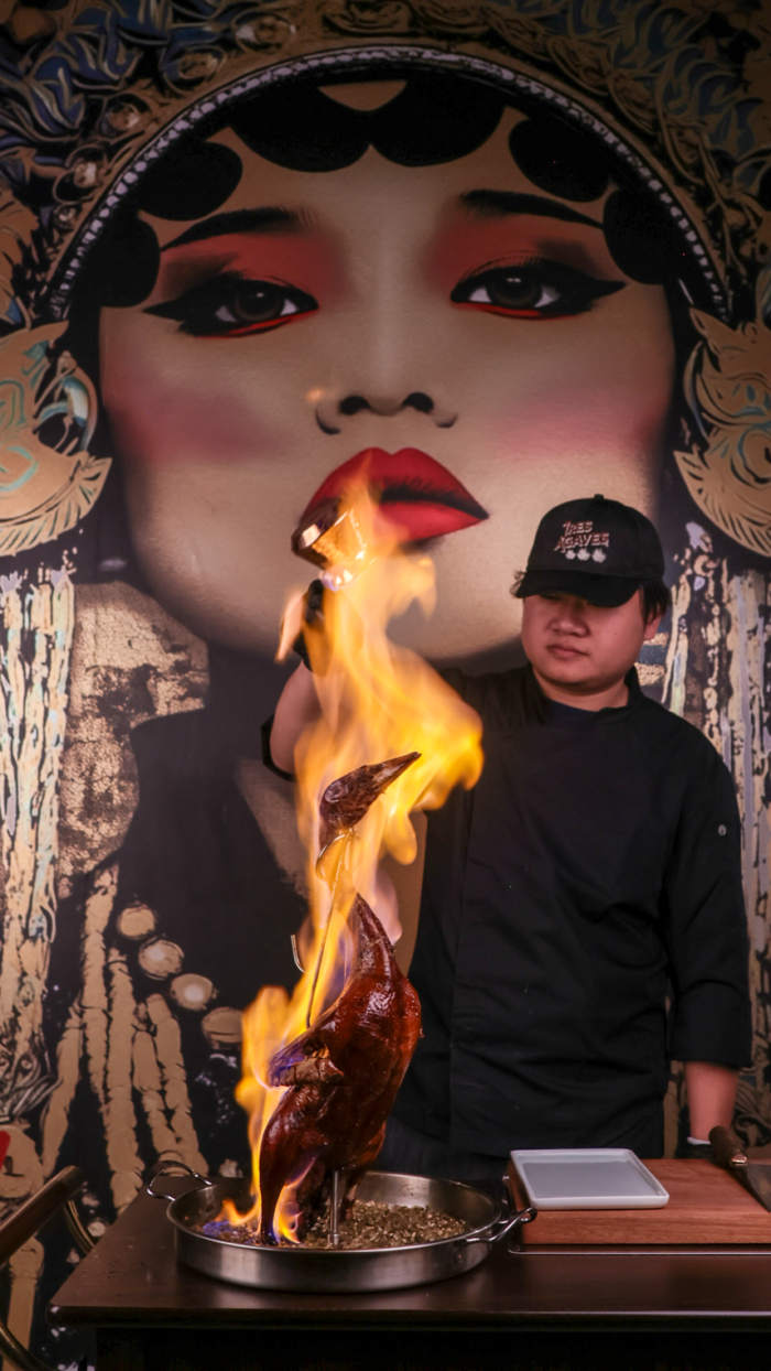A chef in black attire flambés a whole duck, with tall flames rising, in front of a dramatic wall mural of a womans face with bold makeup and traditional headwear.