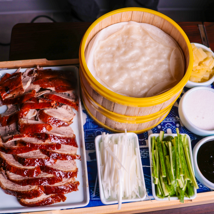 A platter of sliced Peking duck with crispy skin, a bamboo steamer filled with thin pancakes, and small dishes of cucumber, scallions, pineapple, sugar, and hoisin sauce arranged neatly on a table.