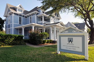 A large, light blue Victorian-style house with white trim sits behind a manicured lawn and bushes. A white sign in front displays text and a logo, with a tree providing shade on the right side.