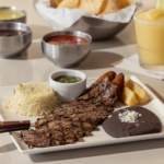 A plate with grilled steak, rice, black beans topped with cheese, fried plantains, yuca fries, green sauce, and a knife. In the background are salsa bowls, tortilla chips, and a yellow frozen drink with a lime.