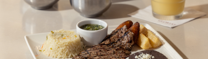 A plate with grilled steak, rice, black beans topped with cheese, fried plantains, yuca fries, green sauce, and a knife. In the background are salsa bowls, tortilla chips, and a yellow frozen drink with a lime.