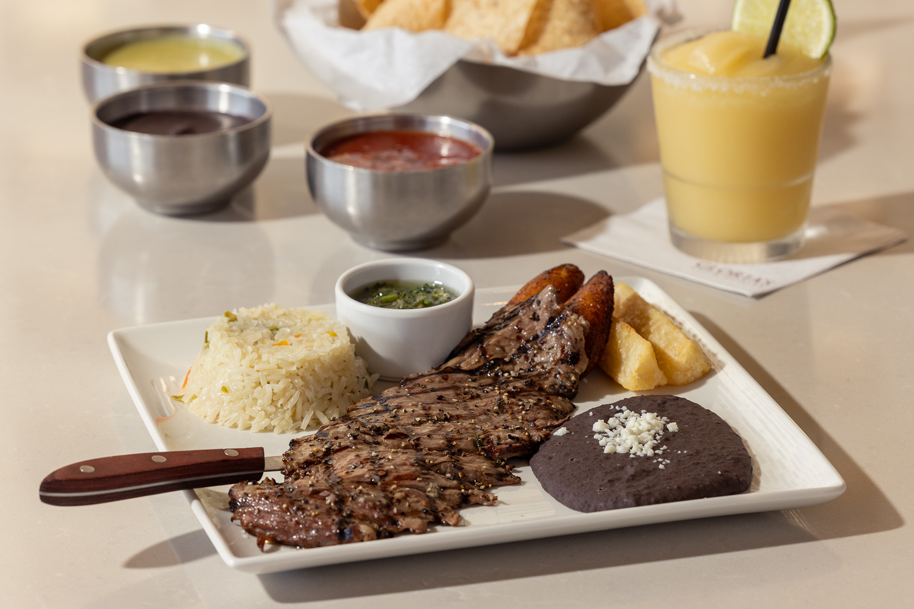 A plate with grilled steak, rice, black beans topped with cheese, fried plantains, yuca fries, green sauce, and a knife. In the background are salsa bowls, tortilla chips, and a yellow frozen drink with a lime.