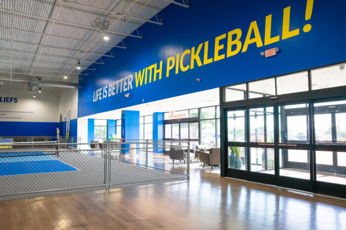 Indoor pickleball court with bright blue walls, large windows, wooden floor, and a bold yellow sign reading LIFE IS BETTER WITH PICKLEBALL! above the court area.