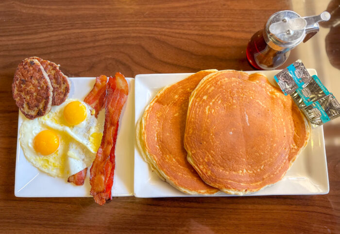 Enjoy a classic Breakfast in Plano: two fried eggs, two sausage patties, crispy bacon strips, three large pancakes with butter packets, and a small jar of syrup—all served on a wooden table.