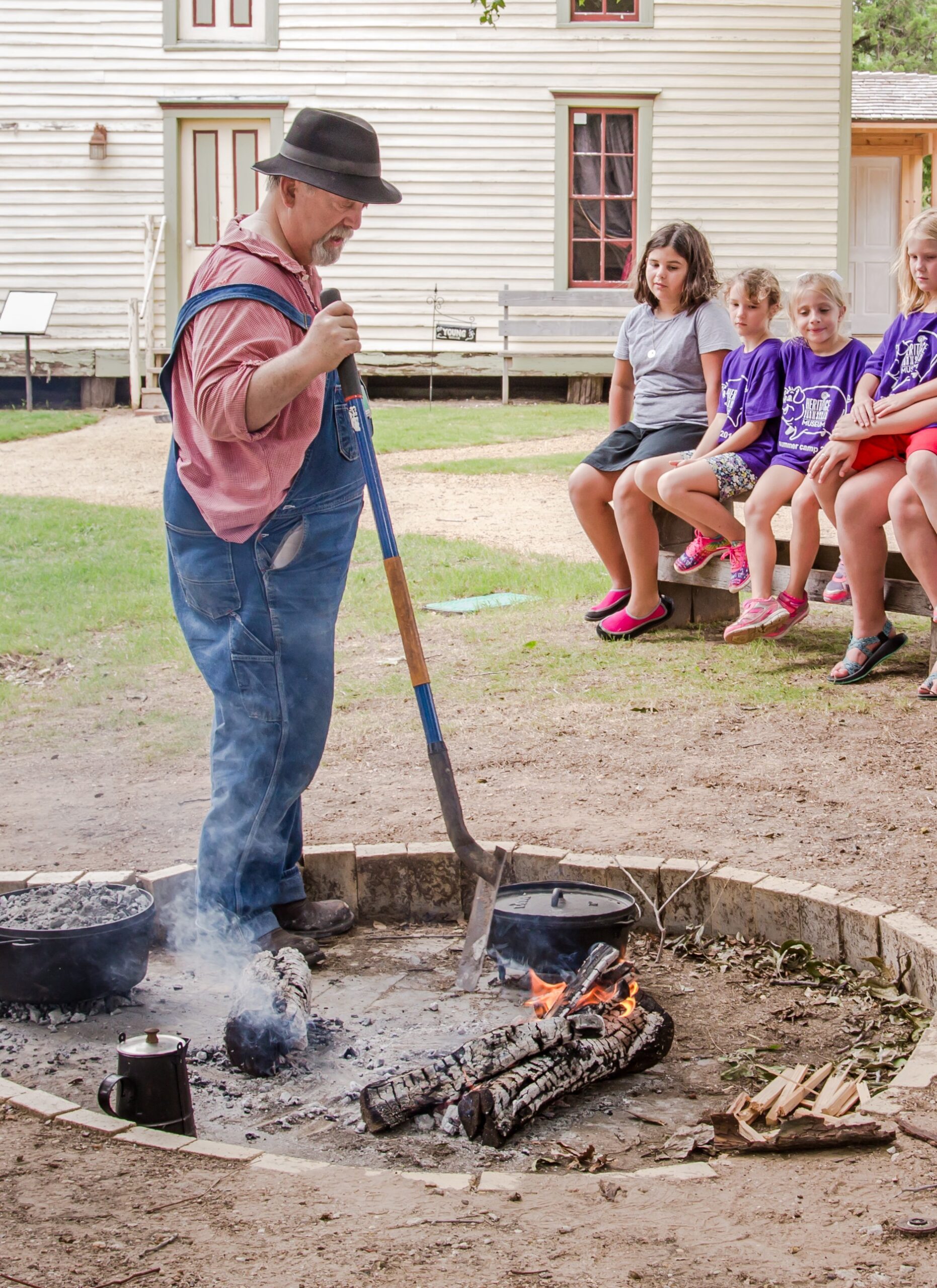A man in overalls and a hat uses a shovel to cook over an outdoor fire pit, while a group of children sit on a bench watching him in front of a wooden building.