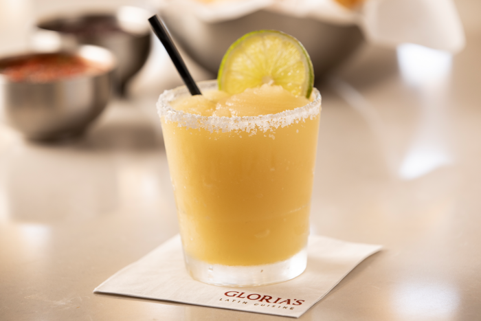 A frozen margarita with a salted rim, lime wheel, and straw is served in a glass on a napkin that reads “GLORIA’S LATIN CUISINE.” Blurred bowls of salsa are visible in the background.