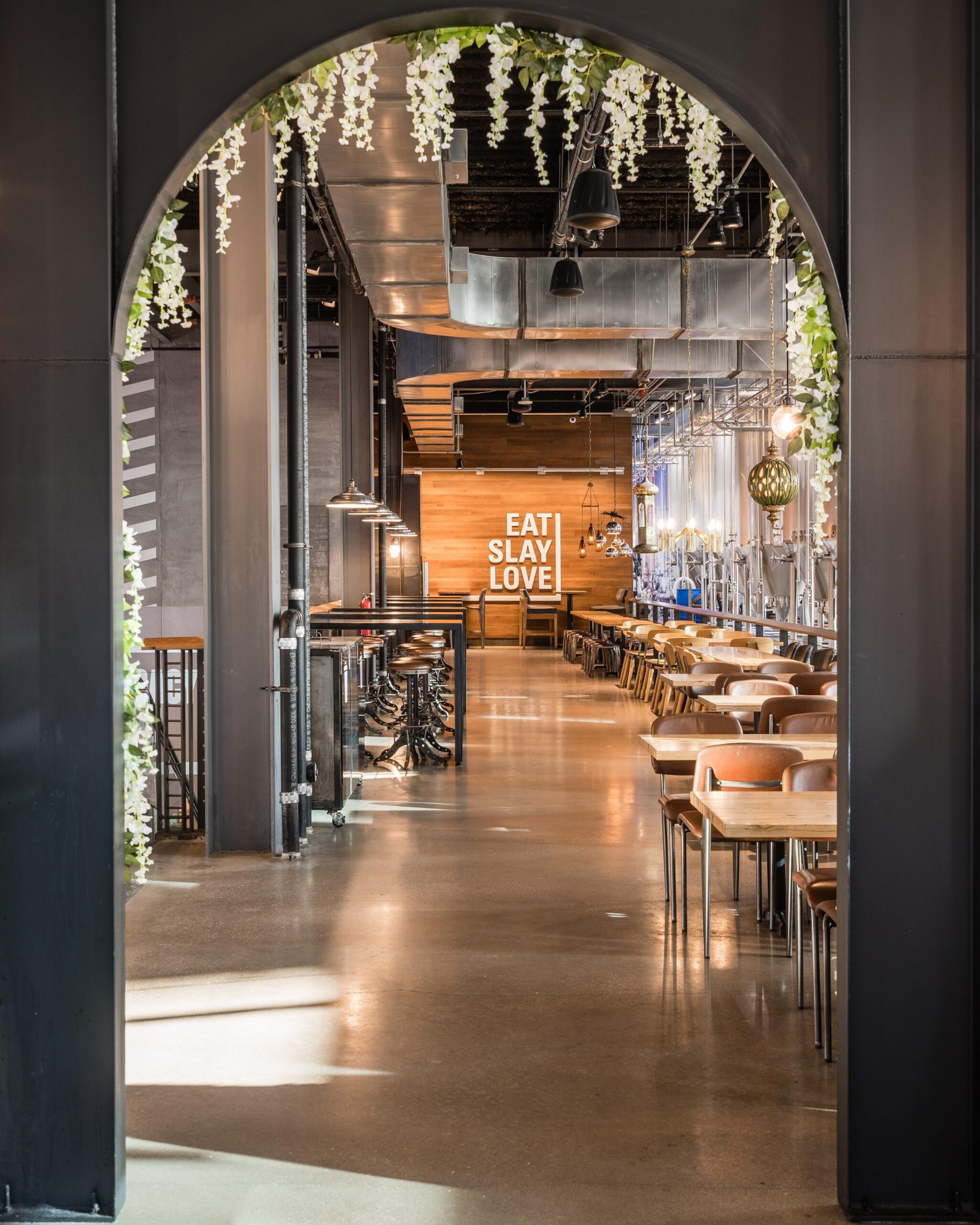 View through an arched entryway into a modern restaurant with wooden tables and chairs, greenery hanging from the ceiling, and a sign at the back wall that reads EAT SLAY LOVE.