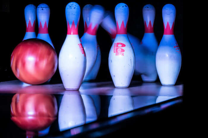 A glowing red bowling ball strikes a set of white bowling pins with red stripes, some of which are beginning to tip over, reflecting on the shiny lane surface.