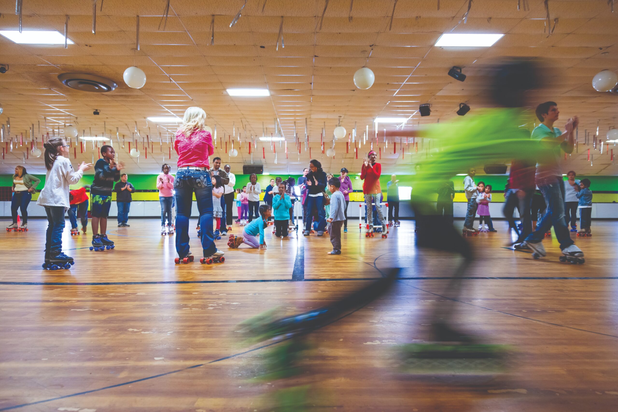 A busy indoor roller skating rink filled with children and adults skating, socializing, and having fun. One skater in bright green is in motion, appearing blurred in the foreground.