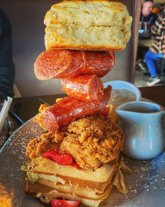 Enjoy Breakfast in Plano with a plate featuring a stack of waffles and fried chicken, sliced sausage skewers, a biscuit on top, fresh strawberries, and a side of white gravy in a small pitcher.