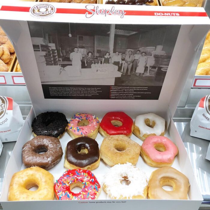 A box of twelve assorted Shipley Do-Nuts—perfect for breakfast in Plano—including glazed, chocolate, sprinkled, coconut, and other frosted varieties. The box lid displays a black-and-white vintage photo of the bakery staff.