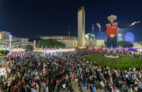 A large crowd gathers at night around a tall cowboy statue in a red shirt and hat at a brightly lit fairground, where food stalls, a Ferris wheel, and festive soccer fans add to the lively atmosphere.