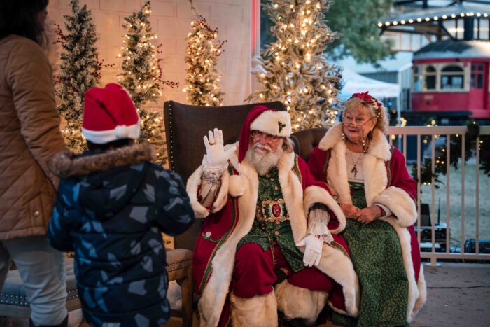 Santa Claus waves to a child wearing a Santa hat, while Mrs. Claus sits beside him. Both are dressed in festive red and green outfits, surrounded by decorated, lit Christmas trees outdoors.