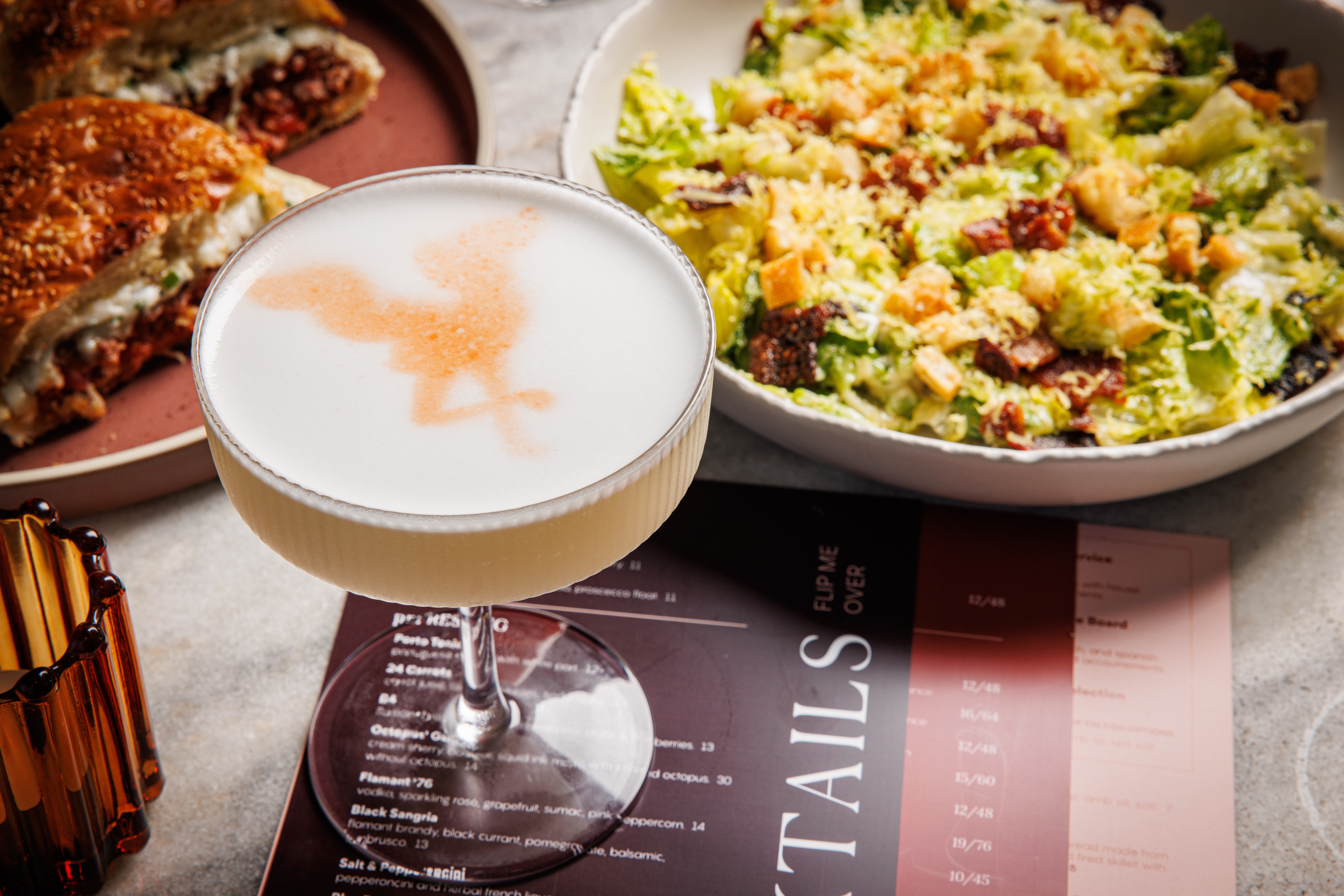 A cocktail with a rooster design in its foam sits on a table alongside a salad, a sandwich on a pink plate, and a cocktail menu—perfect for Group Experiences. The table also holds a brown glass and cutlery.