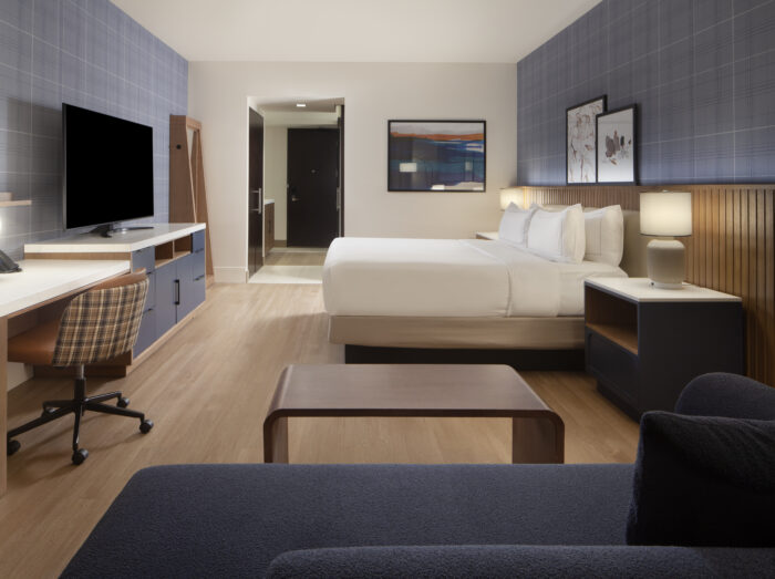 Modern hotel room with a large bed, wall-mounted TV, desk and chair, sofa, wooden floors, two nightstands with lamps, and framed artwork on blue patterned walls. Doorway visible in the background.