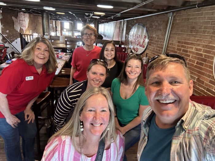 A group of seven smiling adults pose for a selfie inside a rustic restaurant with brick walls and wooden tables. The group includes six women and one man, all wearing casual clothes.