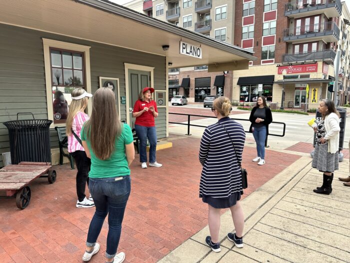A group of people listen to a woman in a red shirt speaking outside a historic train station building with a PLANO sign and apartments and shops visible in the background.