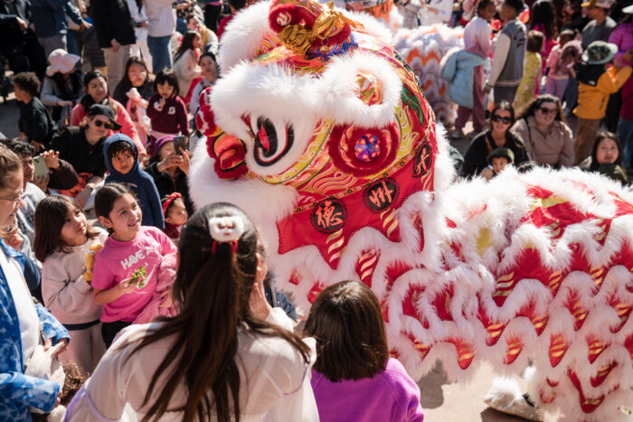 Children and adults gather around a vibrant, colorful lion costume during a festival or parade, smiling and interacting with the performers. The scene is lively, festive, and filled with excitement.